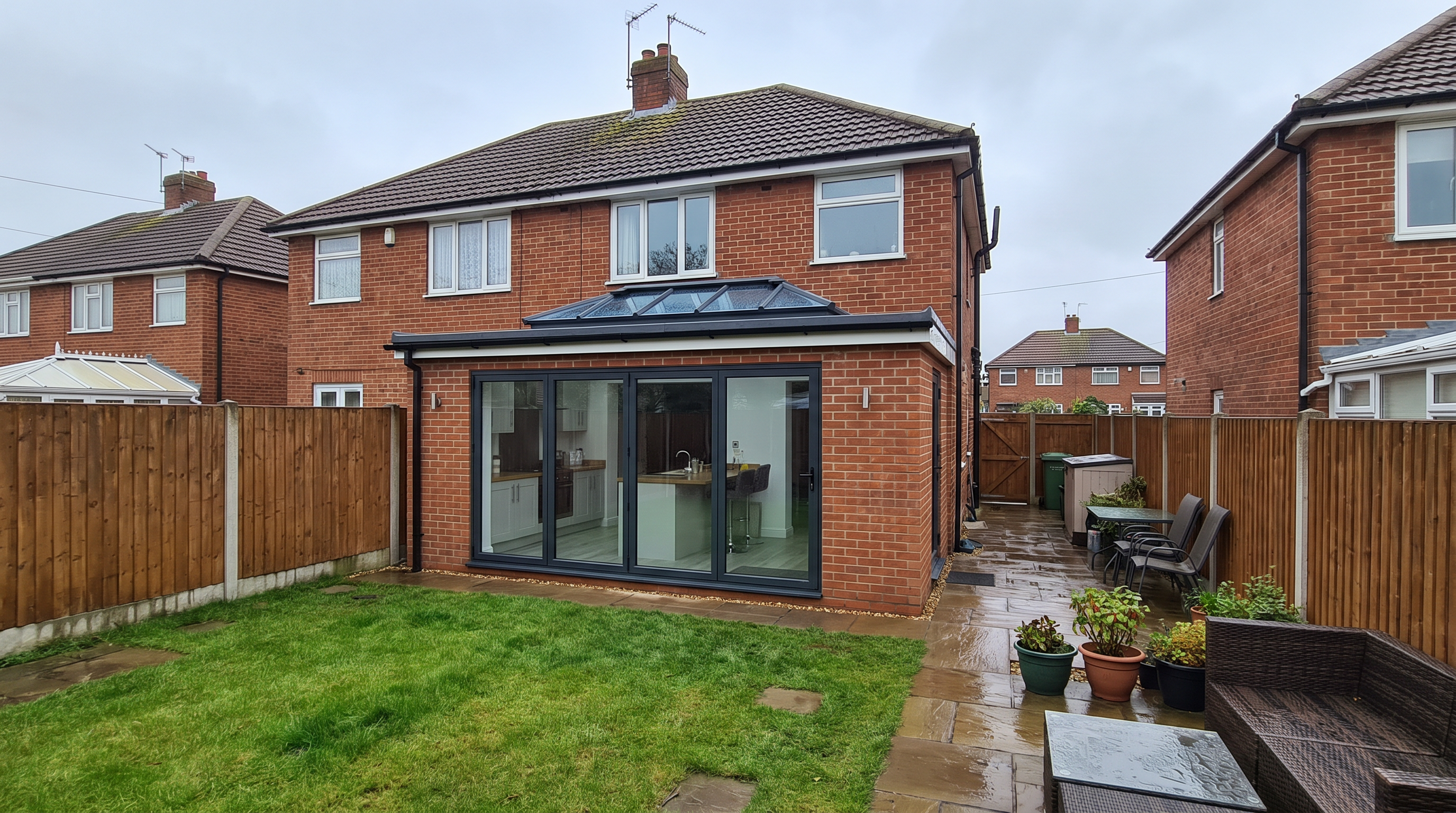 Single-storey rear house extension in brick and timber cladding, its full-width bifold doors open onto a garden patio to reveal a bright open-plan kitchen with rooflights.