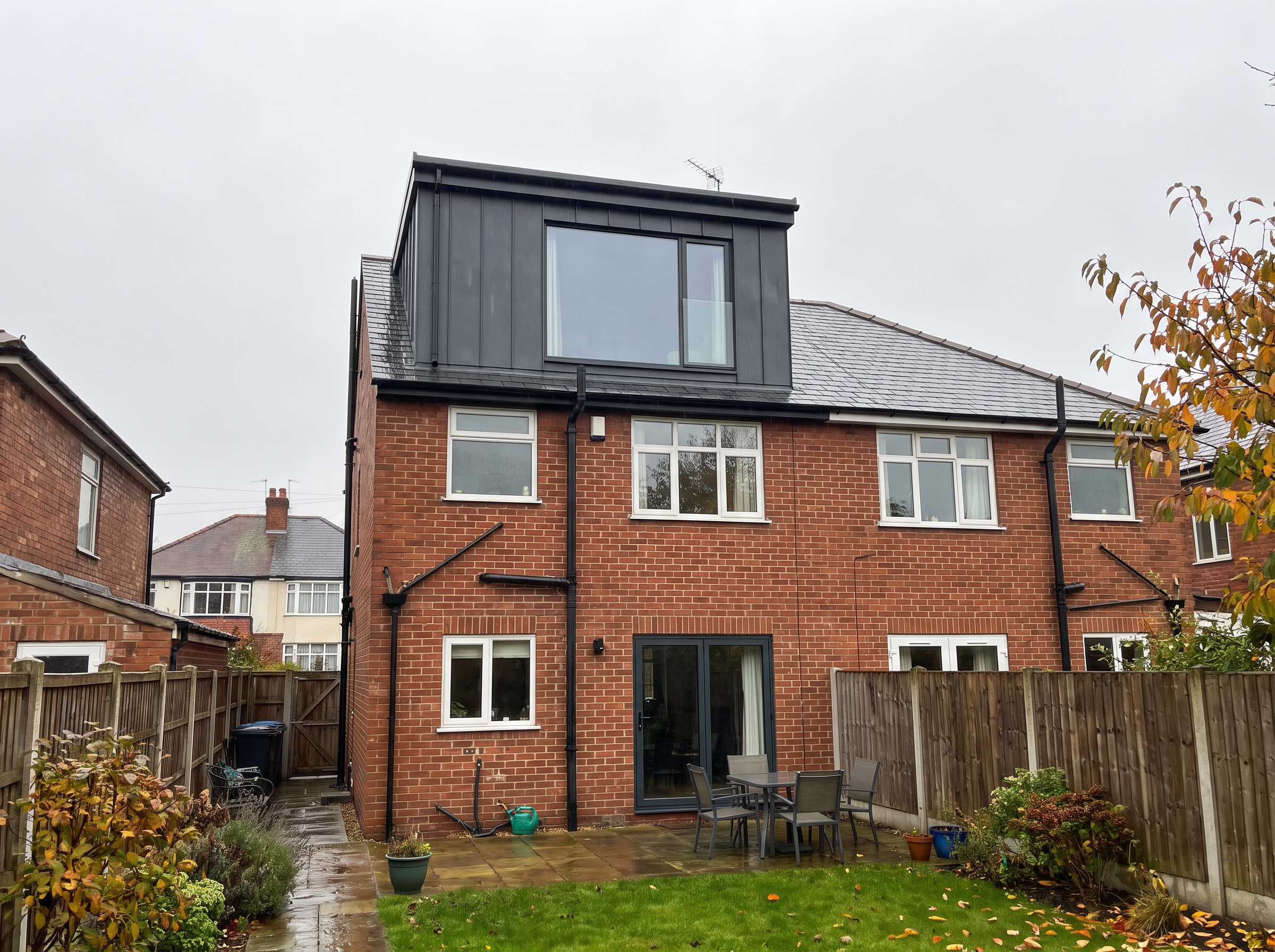 Rear dormer loft conversion clad in dark standing-seam panels with a large picture window, on a red-brick semi-detached house seen from the garden.