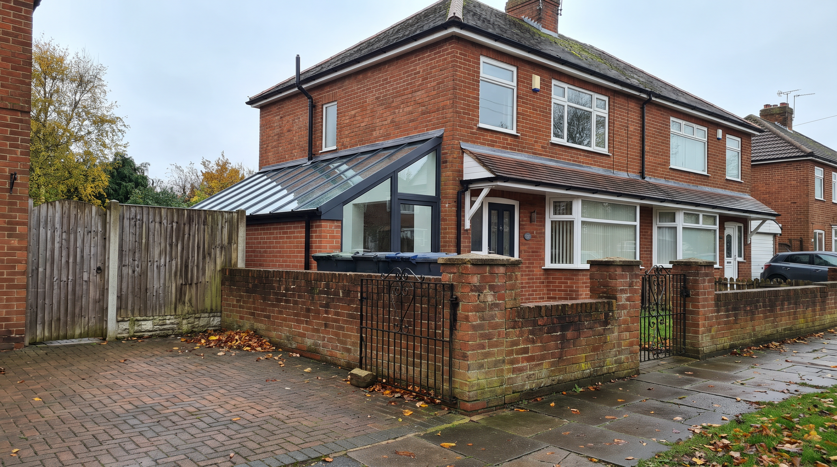 A single-storey side extension with a mono-pitched glazed roof, built alongside a semi-detached red-brick house with a low brick front wall and block-paved driveway.